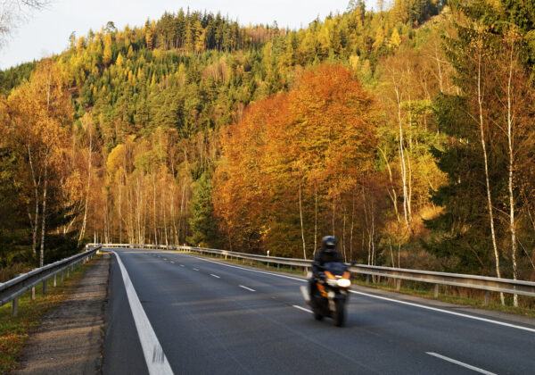 Asphalt road in the autumn landscape with a ride motorcycle, over the road forested mountain, deciduous trees with leaves in bright autumn colors