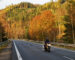 Asphalt road in the autumn landscape with a ride motorcycle, over the road forested mountain, deciduous trees with leaves in bright autumn colors