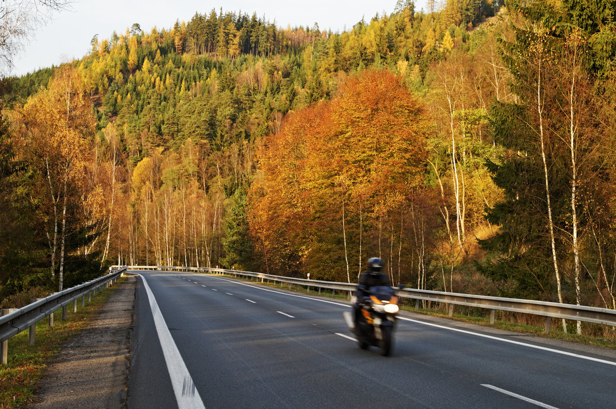 Asphalt road in the autumn landscape with a ride motorcycle, over the road forested mountain, deciduous trees with leaves in bright autumn colors