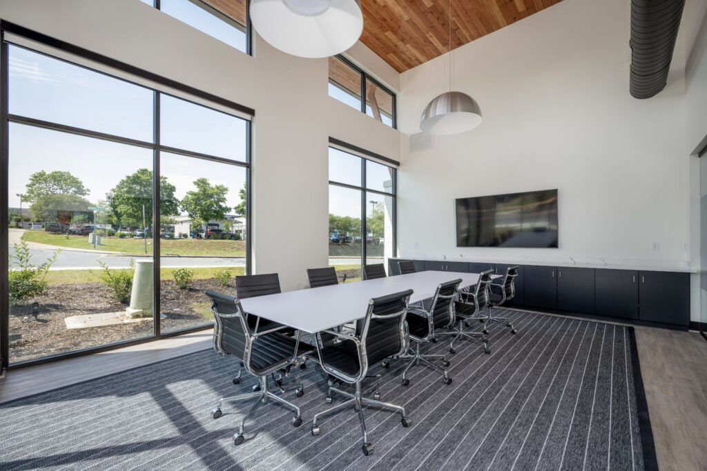Modern meeting room with white conference table, black chairs, wall-mounted TV, and large windows with parking lot views.