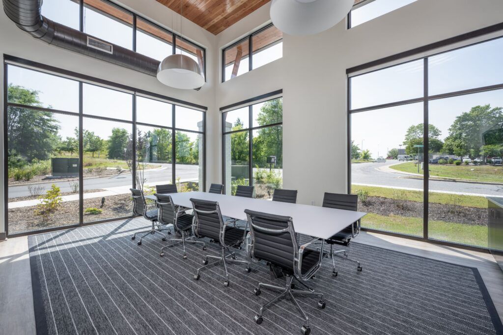 Modern conference room with white table, black chairs, wooden ceiling, and floor-to-ceiling windows overlooking parking lot.