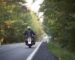 Back view of motorcyclist in black leather jacket and white helmet riding motorbike along hilly road between tall green trees on sunny summer day.